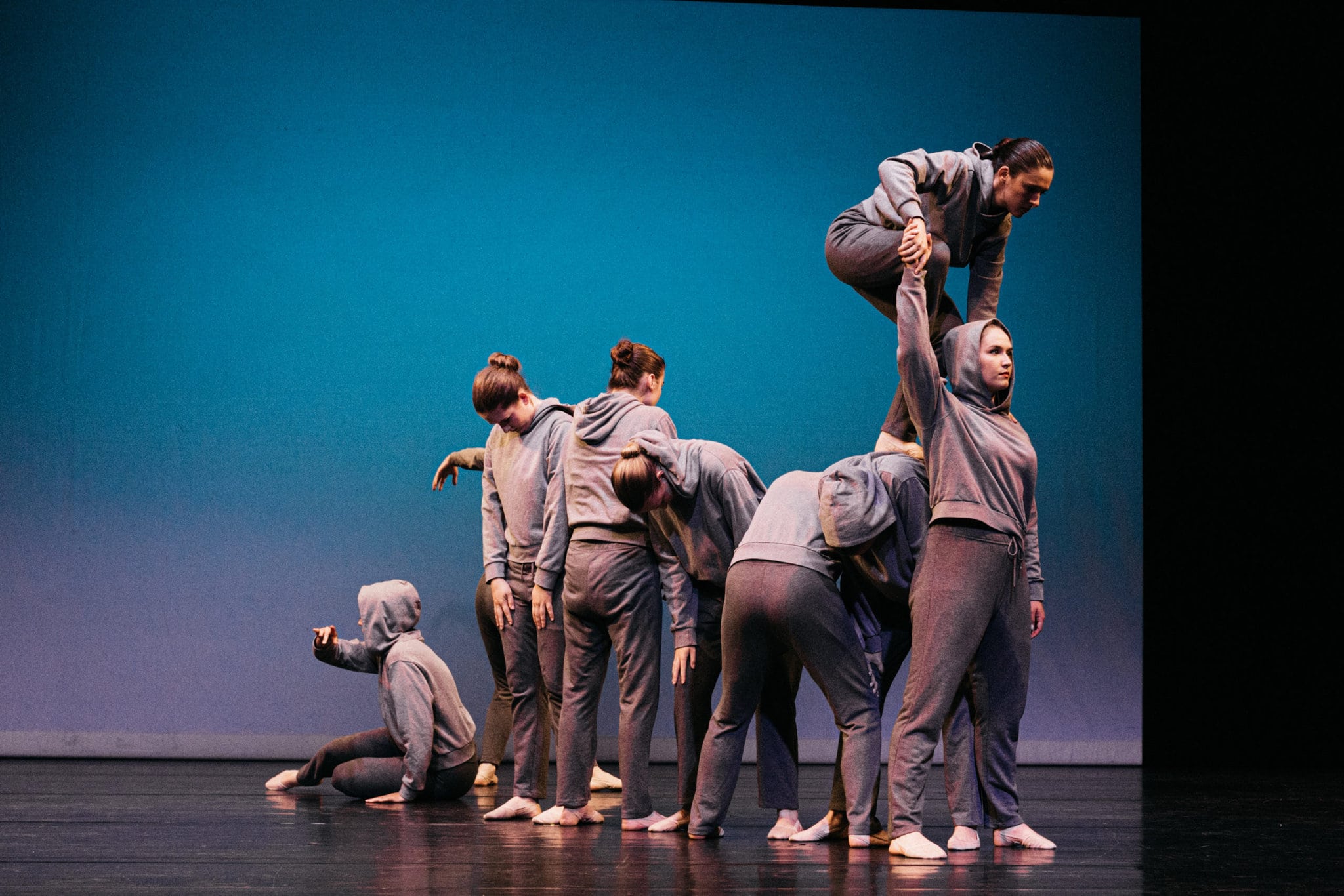 Eine kleine Gruppe junger Menschen steht auf einer Bühne. Sie sind in grau gekleidet und blicken ernst. Sie machen eine Performance.