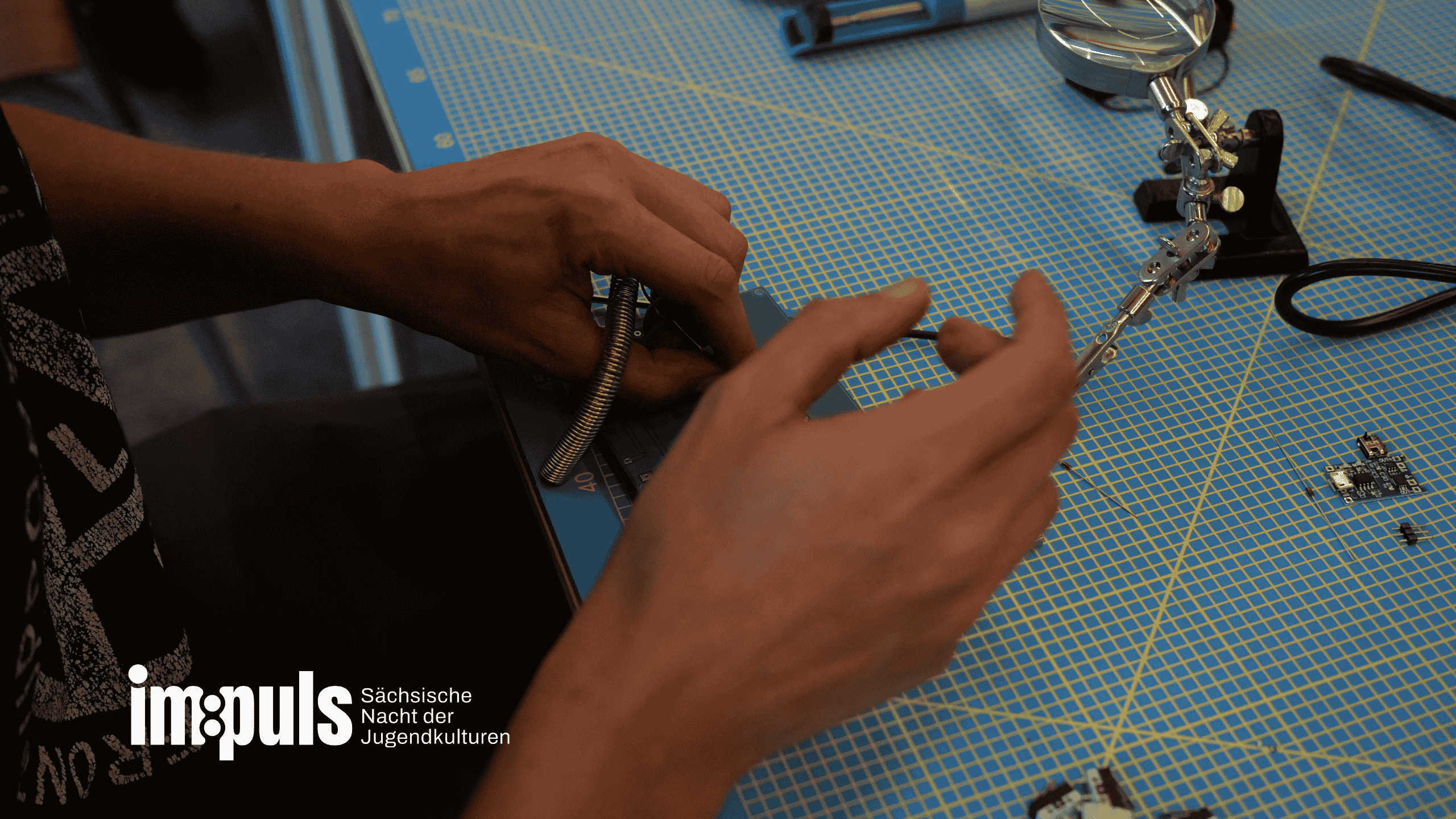 Zwei Hände einer Person sind zu sehen, die etwas bastelt. Auf dem Tisch liegt eine Unterlegmatte, eine Luppe und kleine Platinen