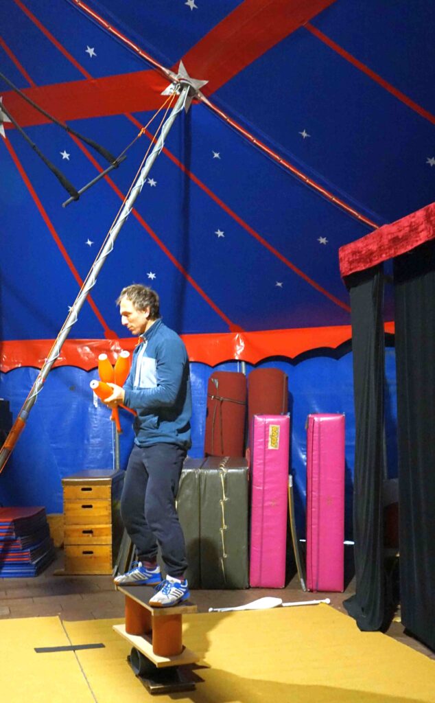 Ein Mann balanciert auf Rollen und Brettern in einem Zirkuszelt. In der Hand hält er Keulen.