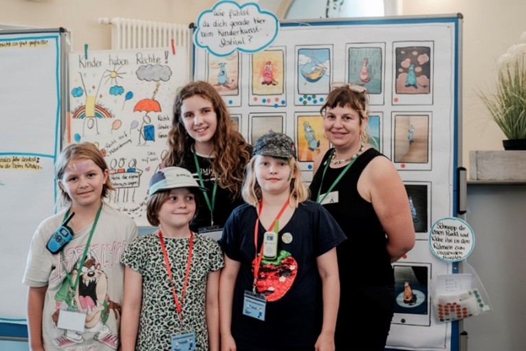 Auf dem Bild sind vier Kinder und eine erwachsene Person zu sehen. Sie lächeln in die Kamera.