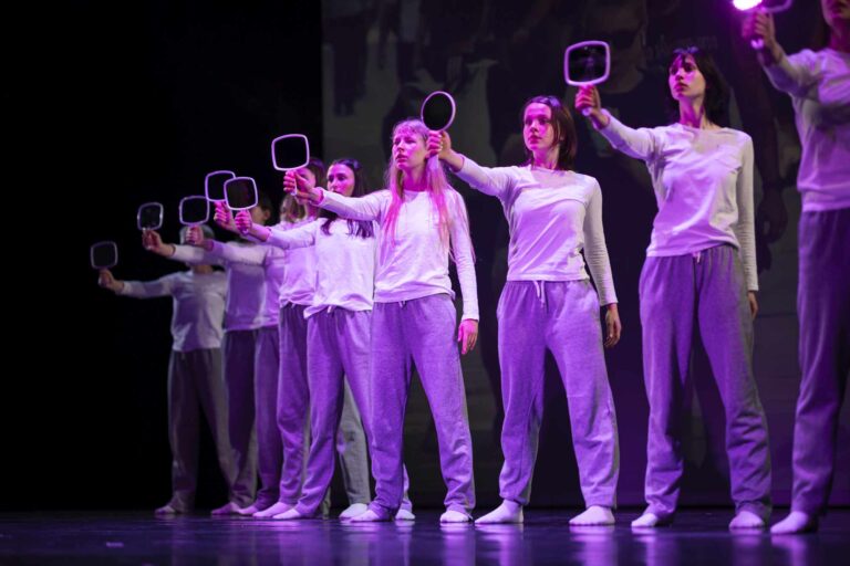 Eine Reihe junger Frauen hält sich auf einer Bühne mit ernstem Ausdruck Spiegel vor das Gesicht, gekleidet in lässiger Sportkleidung.