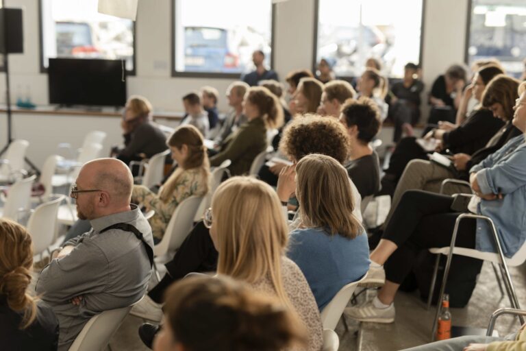 Etwa 30 Personen sitzen in einem Raum und schauen nach vorn.